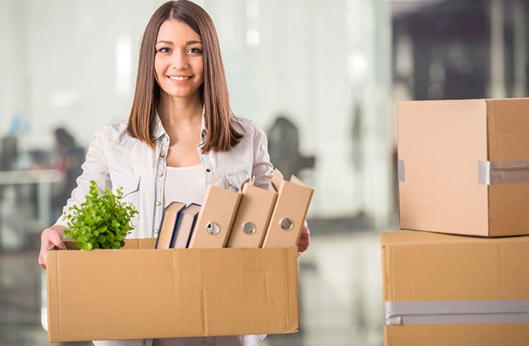 A young female holds a cardboard box with items while standing next to a stack of other cardboard boxes that are filled up and taped closed. 