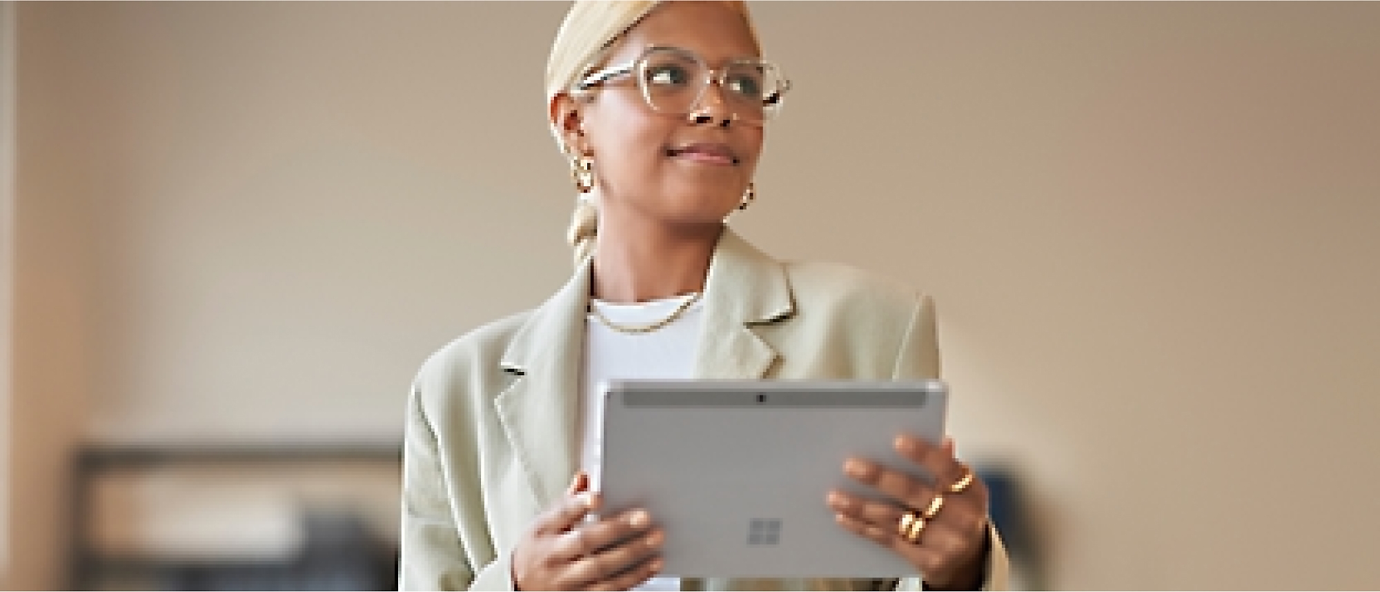 A person with glasses, dressed in professional attire, holds a tablet and looks to the side in a well-lit room.
