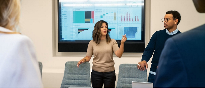 Three people in a meeting room with one person presenting data on a screen.