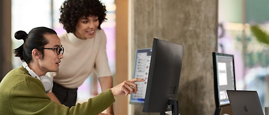 A woman in a white shirt points at a computer screen with a man in glasses and a green sweater watching.