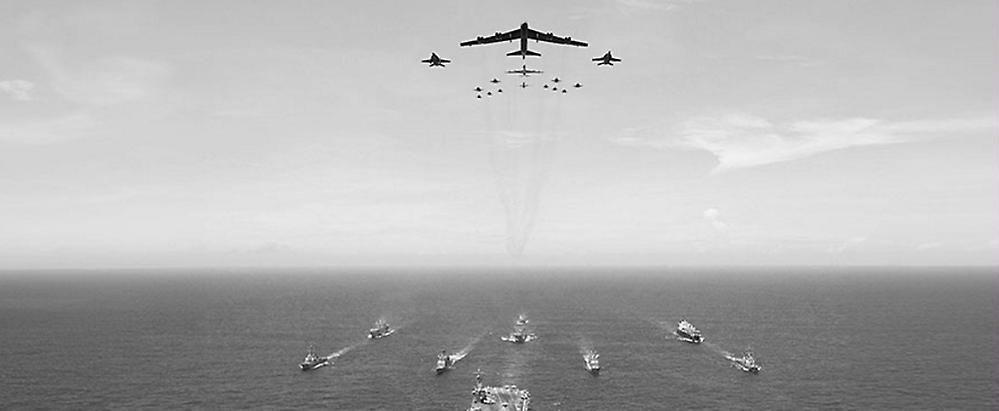Vista aérea de una flota de barcos en el océano con una formación de aviones militar volando sobre el cielo, dejando huellas en el cielo.