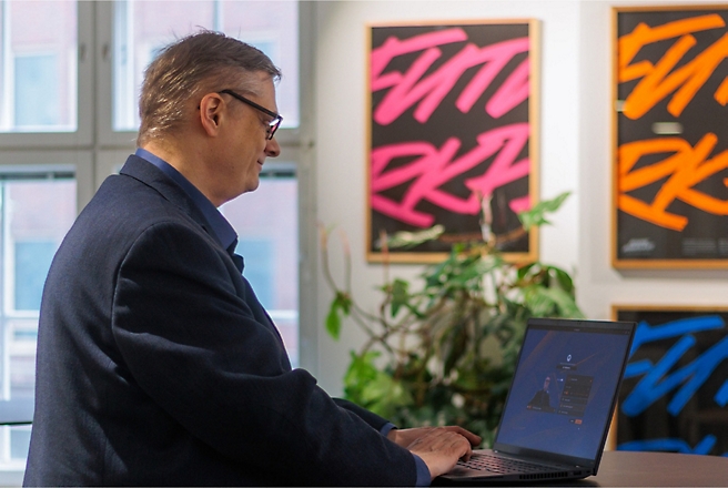 Een persoon in een pak werkt op een laptop in een kantoor met kleurrijk kunstwerken en planten op de achtergrond.
