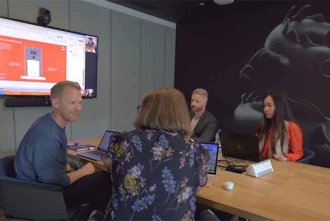 Vier mensen in een vergaderruimte, bezig met een videogesprek en presentatie, met laptops open en een scherm met dia's.