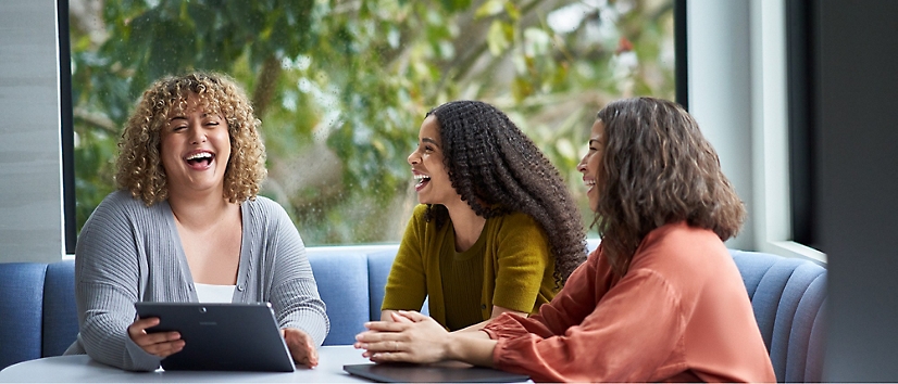 Three people sitting at a table, engaged in conversation and laughing, with a window and greenery in the background.