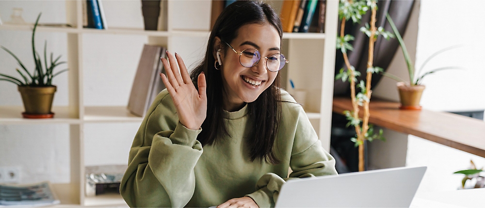 Uma pessoa com suéter verde e óculos senta-se em uma mesa em frente a um notebook, sorrindo e acenando.