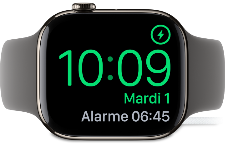 Apple Watch placée sur sa tranche et connectée au chargeur. L’écran montre le symbole de recharge dans le coin supérieur droit, l’heure actuelle en dessous et l’heure de la prochaine alarme.