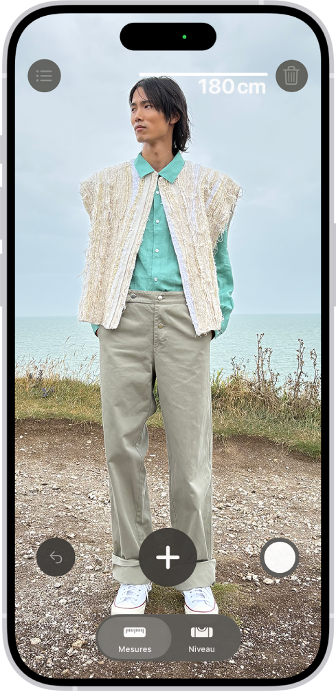 La taille d’une personne en cours de mesure dans l’app Mesure, avec la mesure de la taille affichée au-dessus de la tête de la personne. Le bouton Prendre une photo se trouve vers le coin inférieur droit pour prendre une photo de la mesure. L’indicateur Caméra en cours d’utilisation vert apparaît en haut.