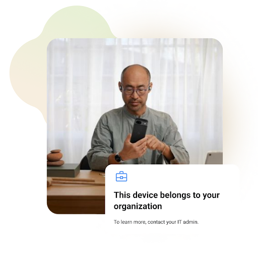 Man using a smartphone while seated at a desk. The setting appears to be a home office or workspace. A message on the screen reads, "This device belongs to your organization. To learn more, contact your IT admin."