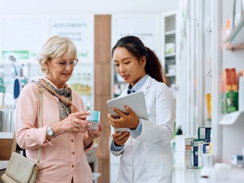 Chinese pharmacist using digital tablet while assisting her senior customer in buying medicine in a pharmacy.