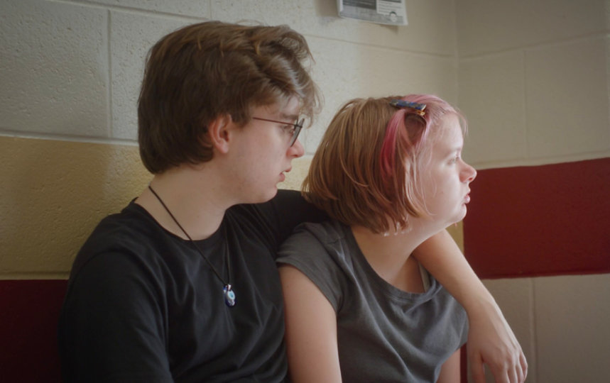 Archer sits with his arm around Della as they look to the right.