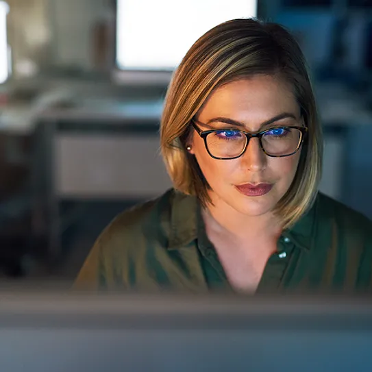 A young woman is sitting at a desk, smiling and writing notes while looking at a computer screen.