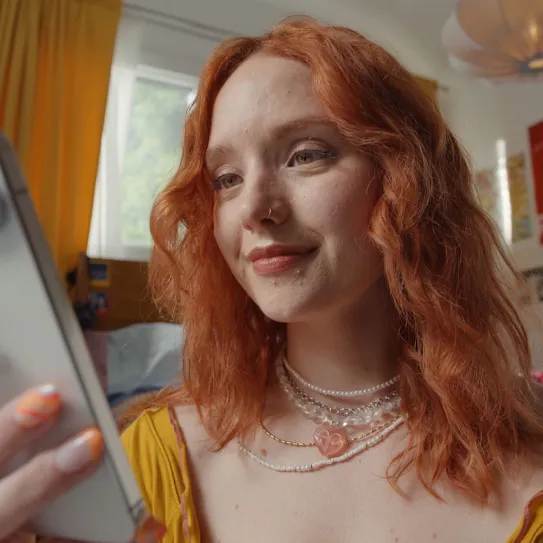 A person with red hair looks at their Pixel phone. They are wearing several necklaces and a yellow shirt.